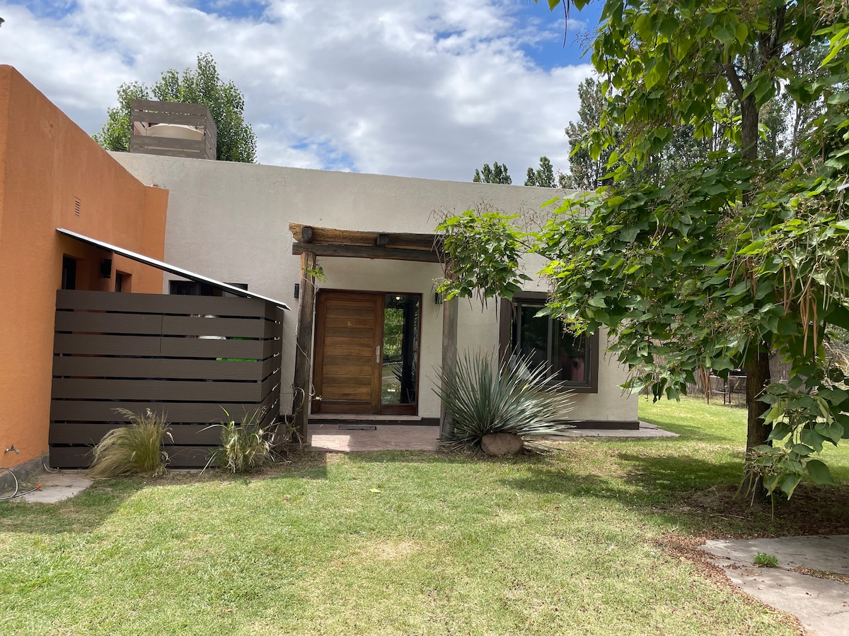 The exterior of a modern building is showcased, featuring a natural stone façade with a wooden door. Lush green grass surrounds the structure, accompanied by trees and shrubs. A shaded area is indicated by a wooden fence, contributing to the overall tranquil atmosphere.
