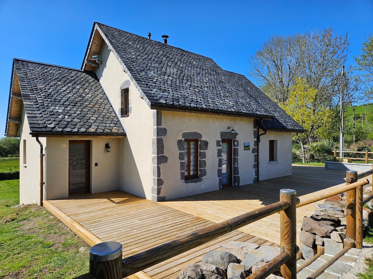 The exterior of a two-story house is presented, featuring a slate roof and light-colored walls. A spacious wooden deck extends from the front, bordered by a wooden railing. Surrounding greenery provides a natural setting, contrasting with the building's architecture.