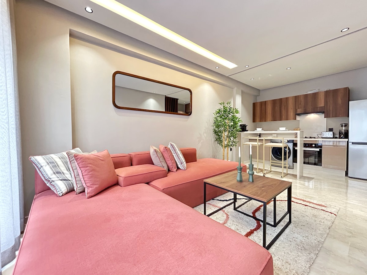 A bright and spacious living area features a large pink sectional sofa with decorative pillows, complemented by a minimalist coffee table. An open-plan kitchen area is visible in the background, equipped with modern appliances and a clean design. Natural light fills the room, enhancing the contemporary ambiance.