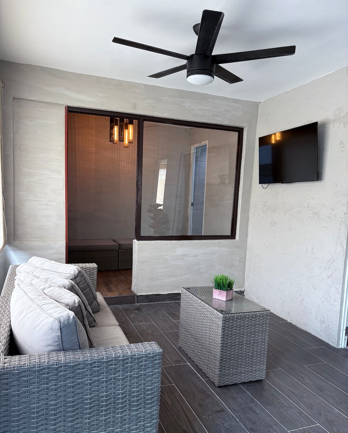 A cozy seating area features a comfortable gray sofa adorned with soft cushions, positioned next to a textured coffee table. A wall-mounted television is visible, and a ceiling fan hangs overhead. Natural light filters through glass doors that lead to an adjoining space.