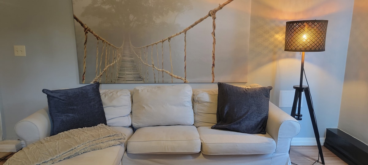 A cozy living area showcases a white sofa adorned with two dark pillows and a light throw blanket. A wall mural of a rope bridge adds a calming backdrop, while a stylish floor lamp provides warm illumination.