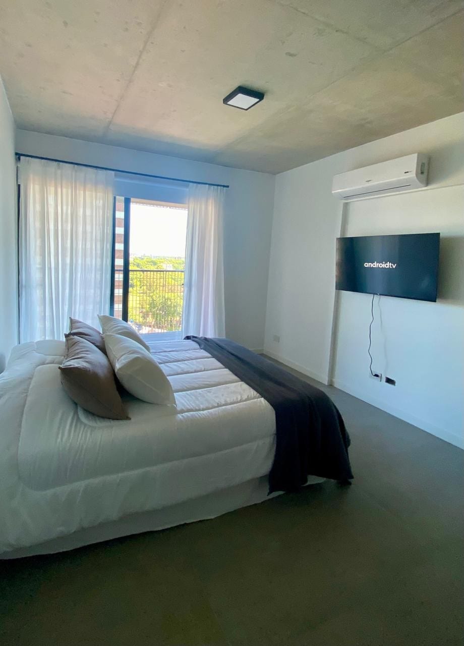 A well-appointed bedroom features a spacious bed with neatly arranged pillows. A television is mounted on the wall, and light filters through sheer curtains, casting a soft glow. A sliding glass door provides access to a balcony, offering views of the surroundings.