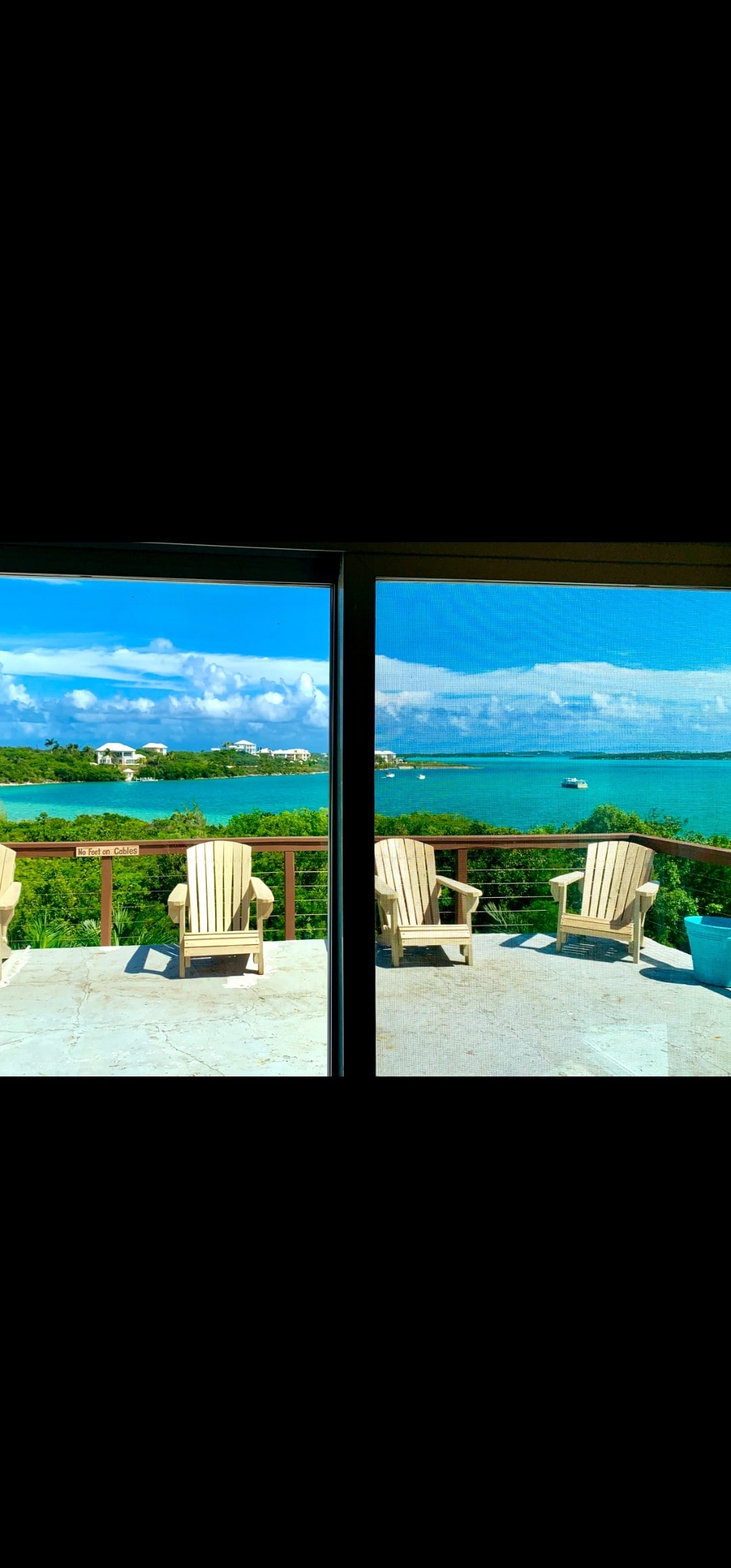 A spacious balcony is seen through large glass doors, featuring several light-colored Adirondack chairs positioned for optimal ocean views. The vibrant blue waters of Elizabeth Harbour are visible, framed by lush greenery and a clear sky, creating a serene outdoor setting.