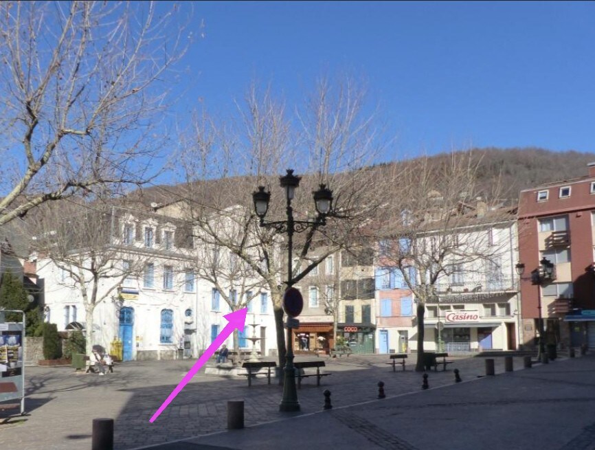 A quiet plaza features bare trees and a streetlamp at the forefront. Nearby buildings exhibit a mix of colors and clear architectural lines. Shops, including a casino, are visible in the background, with a street sign and pedestrian-friendly layout enhancing accessibility.