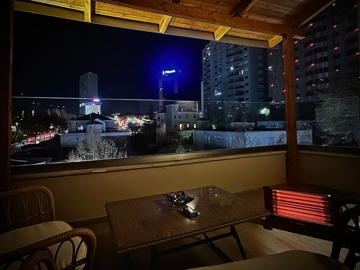A private balcony is illuminated at night, offering a view of the vibrant city lights and buildings in the distance. A cozy seating arrangement features a table and chairs, complemented by a heater to provide warmth during cooler evenings.