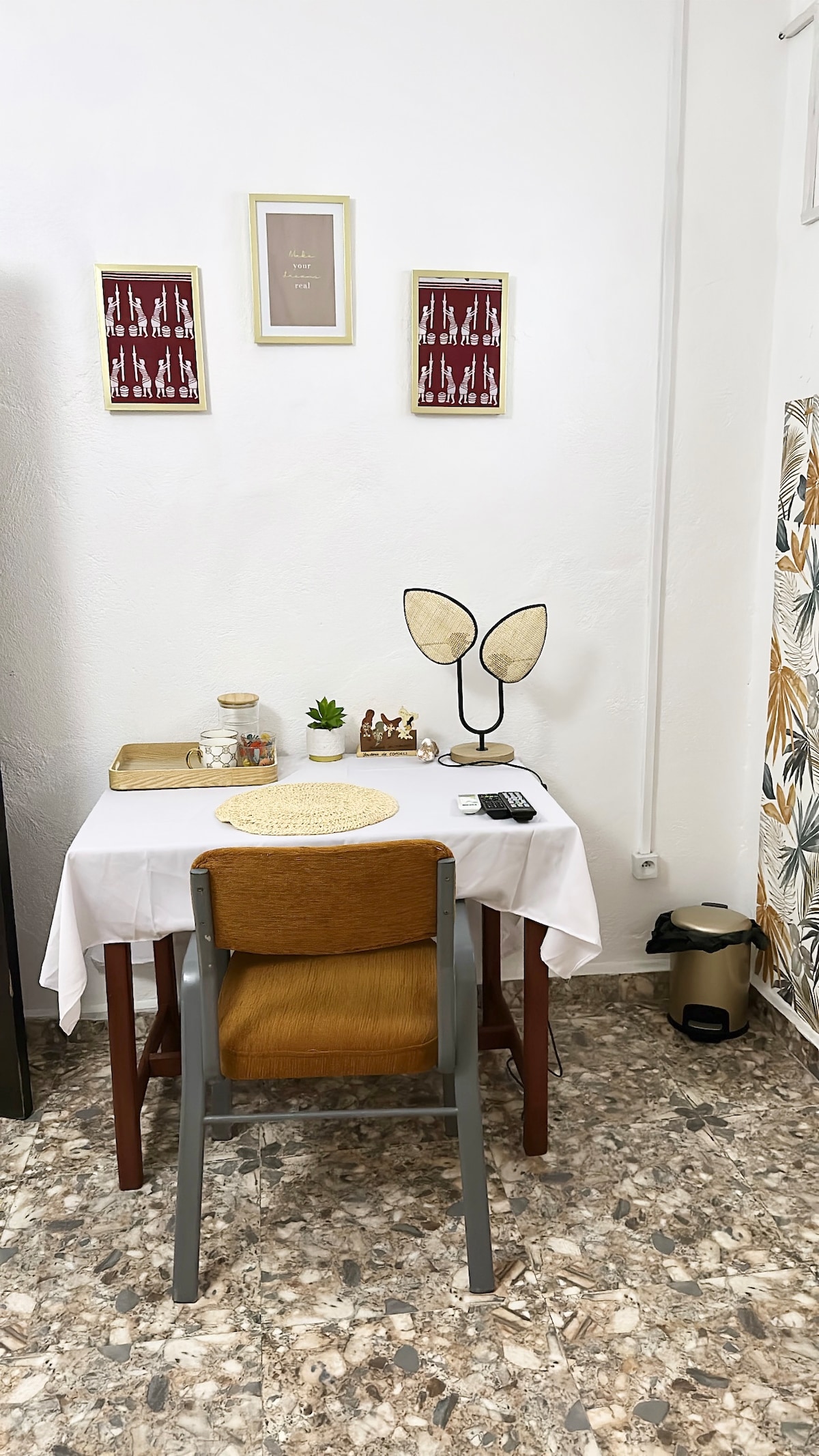 A simple workspace is presented with a wooden chair and a table draped in a white cloth. Decorative items, including a small plant and framed artwork, adorn the walls. A lamp with a leaf design and a wastebasket are included, creating a functional and organized area.