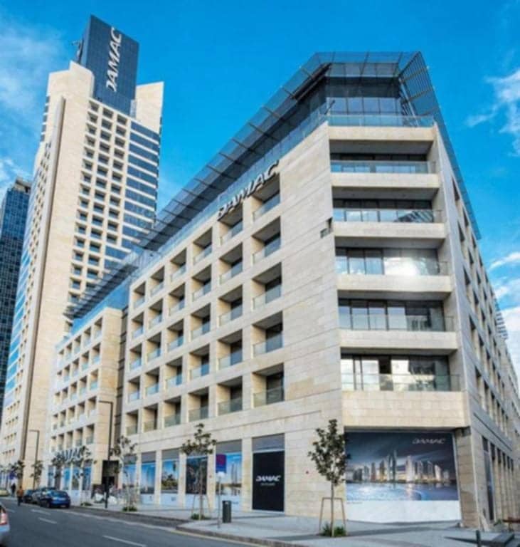 The exterior of a modern building is showcased, characterized by angular architecture and large glass windows. The façade features a combination of stone and glass, with trees lining the street at the base, contributing to a vibrant urban scene.