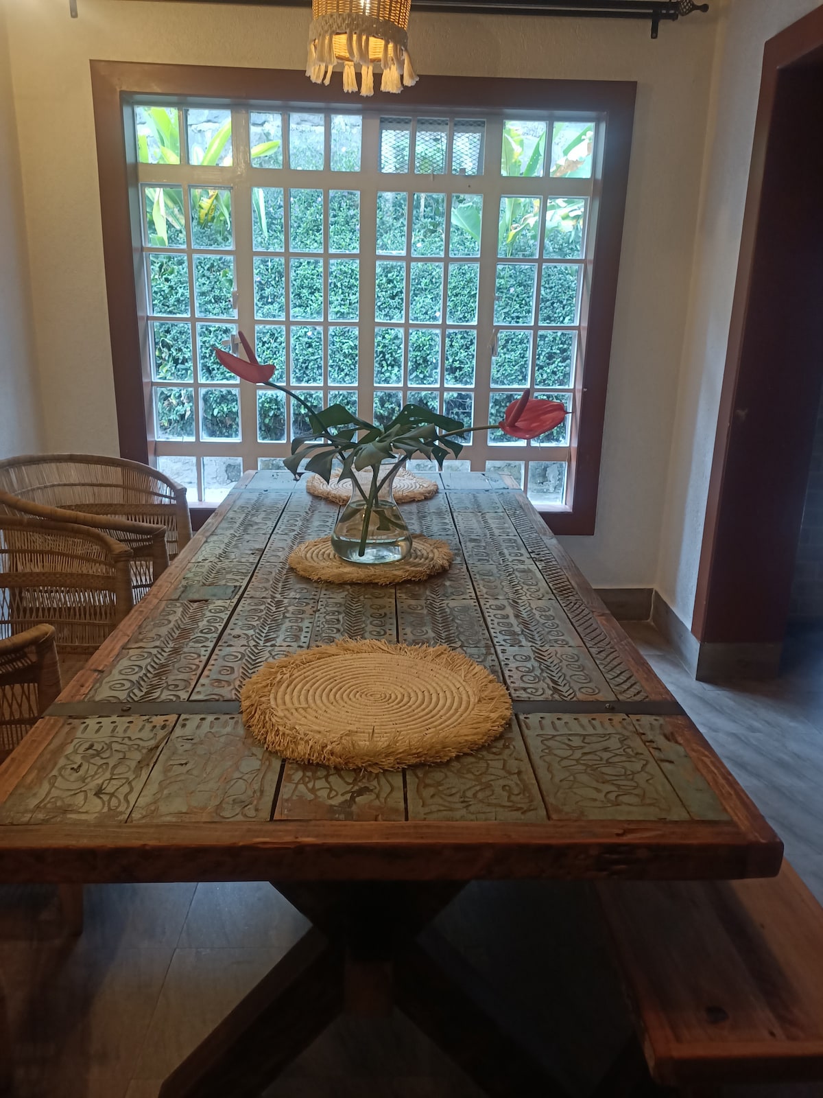 A spacious dining area features a large wooden table adorned with intricate carvings. Natural light enters through a grid of windows, illuminating the surface. Four woven chairs are positioned around the table, complemented by decorative placemats and a small plant centerpiece.