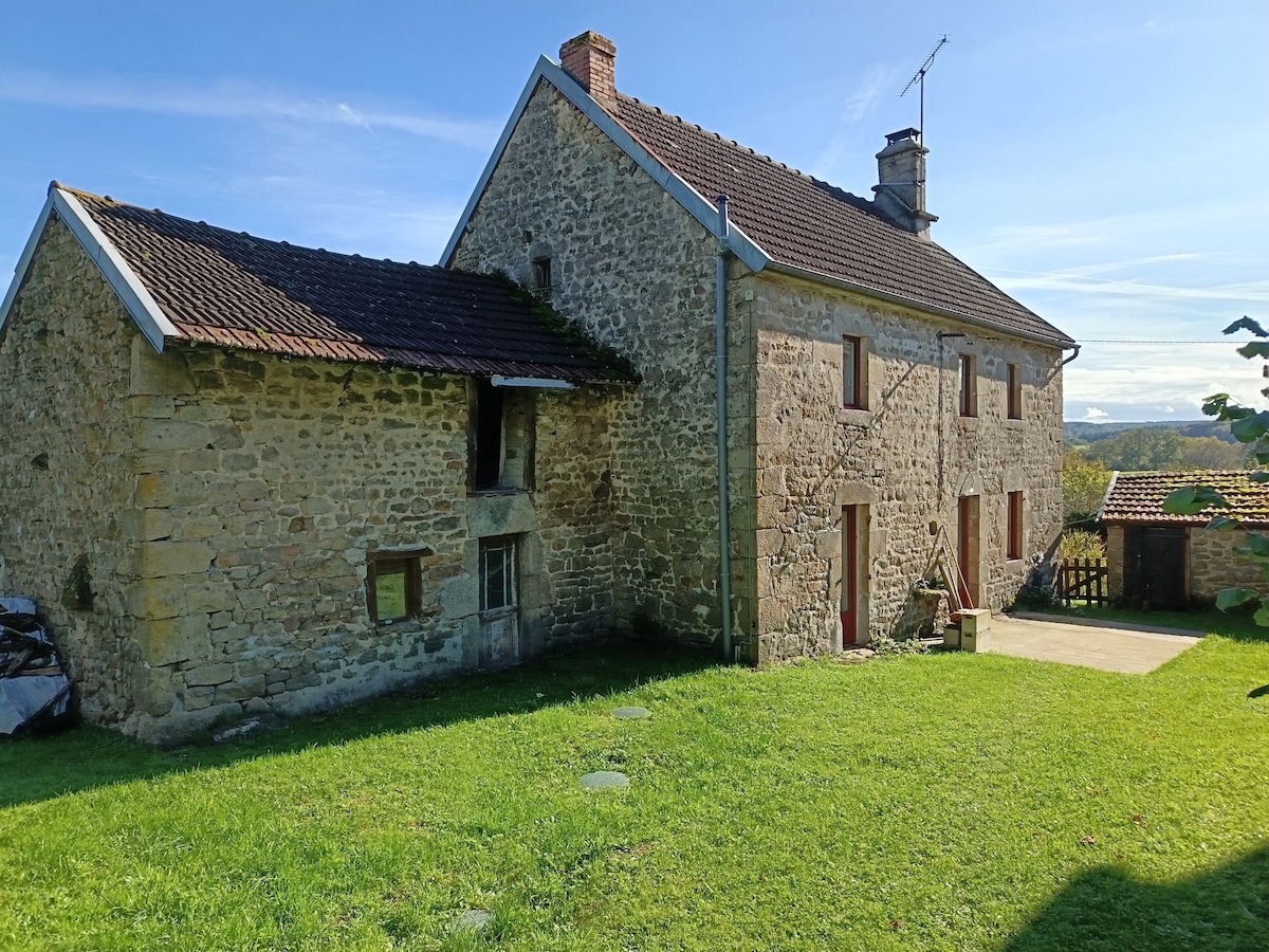 The exterior of a charming stone house is presented, featuring a sloped roof and multiple windows. A well-maintained lawn surrounds the structure, with a paved area visible in front. Scenic views of the countryside are seen in the background under a clear blue sky.