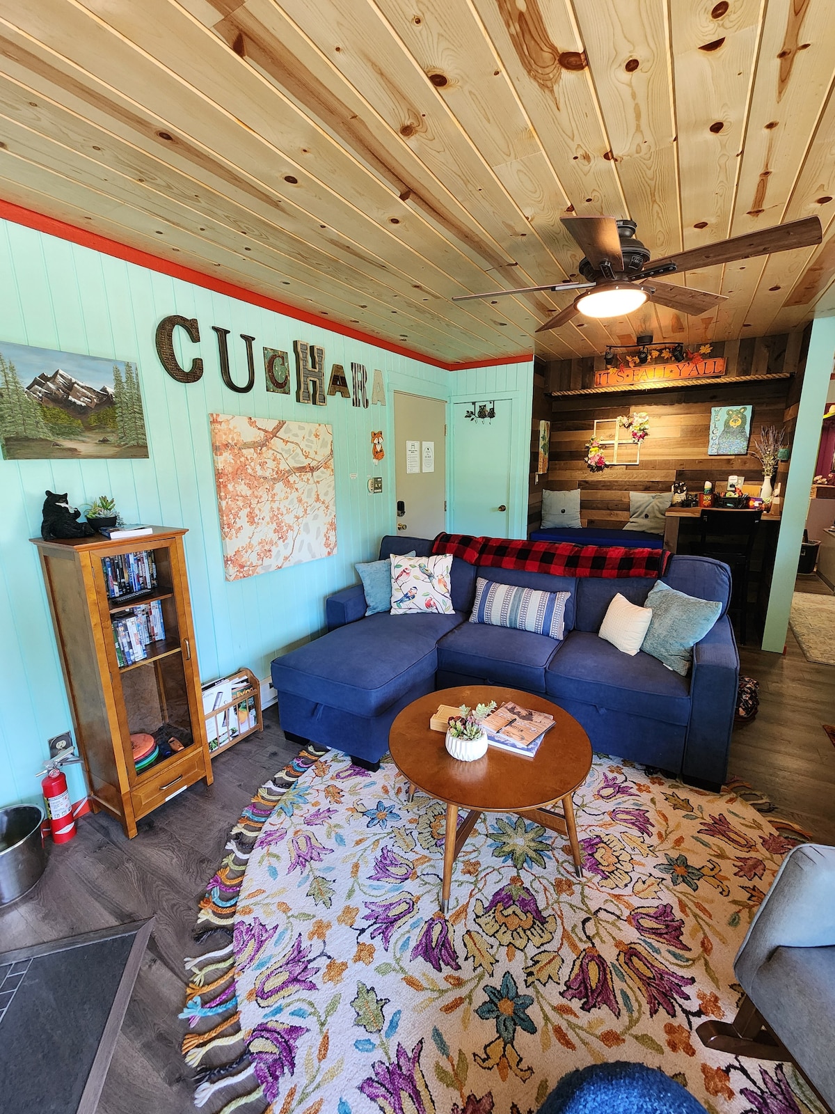 A cozy living area features a blue chaise couch adorned with colorful throw pillows. A round coffee table sits on a decorative rug, with a collection of books and magazines neatly arranged. The walls display warm wood paneling and inviting artwork, contributing to a relaxing atmosphere.