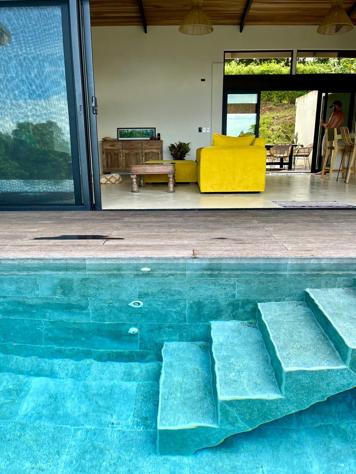 A clear view of an infinity pool, featuring stone steps leading into the water. In the background, large glass doors open to a bright living space with a yellow accent chair and wooden coffee table, offering a seamless connection between indoor and outdoor areas.