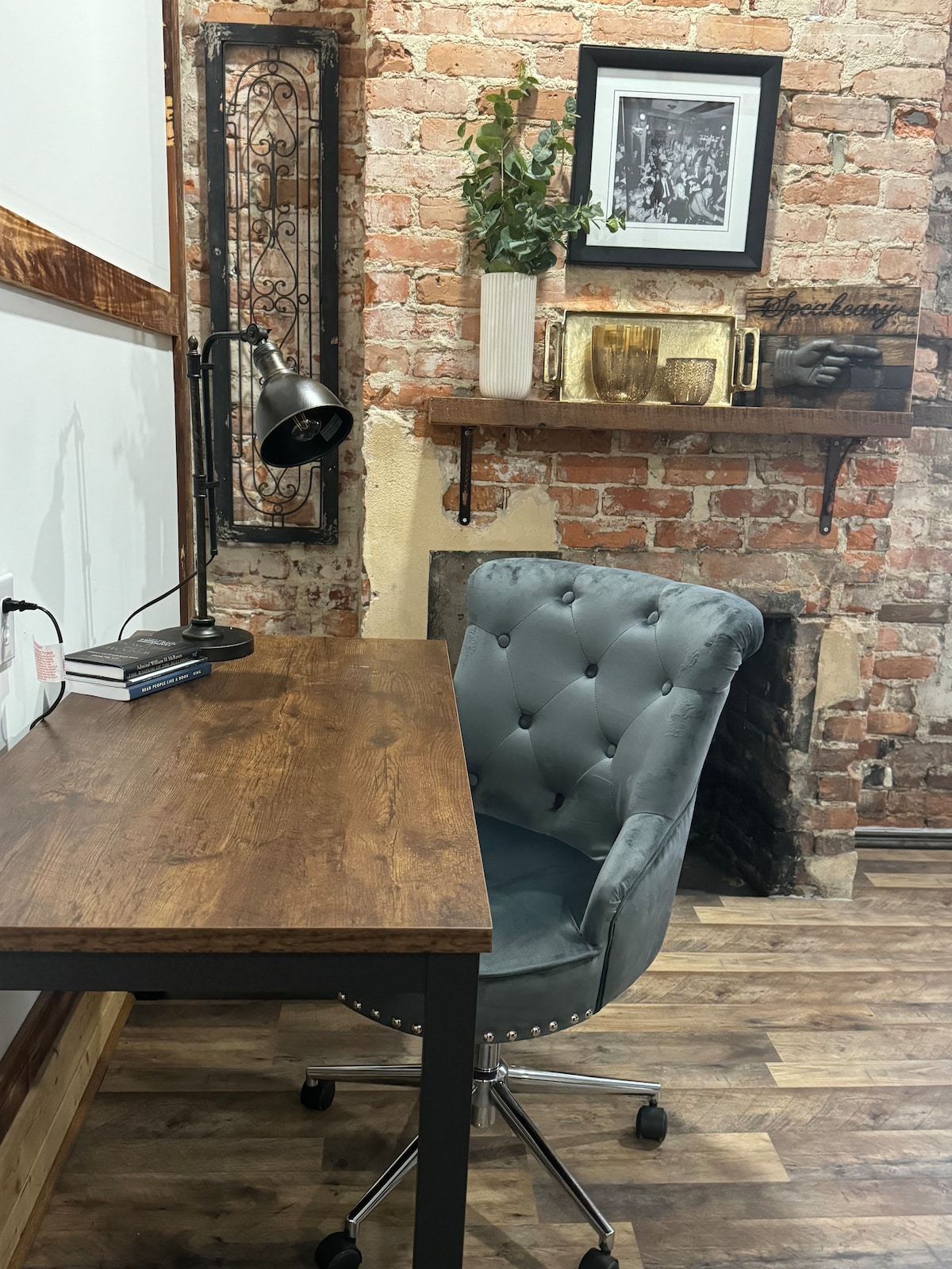 A workspace is presented with a rustic wooden desk and a plush gray chair with button-tufted upholstery. A black desk lamp provides focused lighting. The wall features exposed brick and a decorative shelf holding plants and decorative items, enhancing the ambiance.