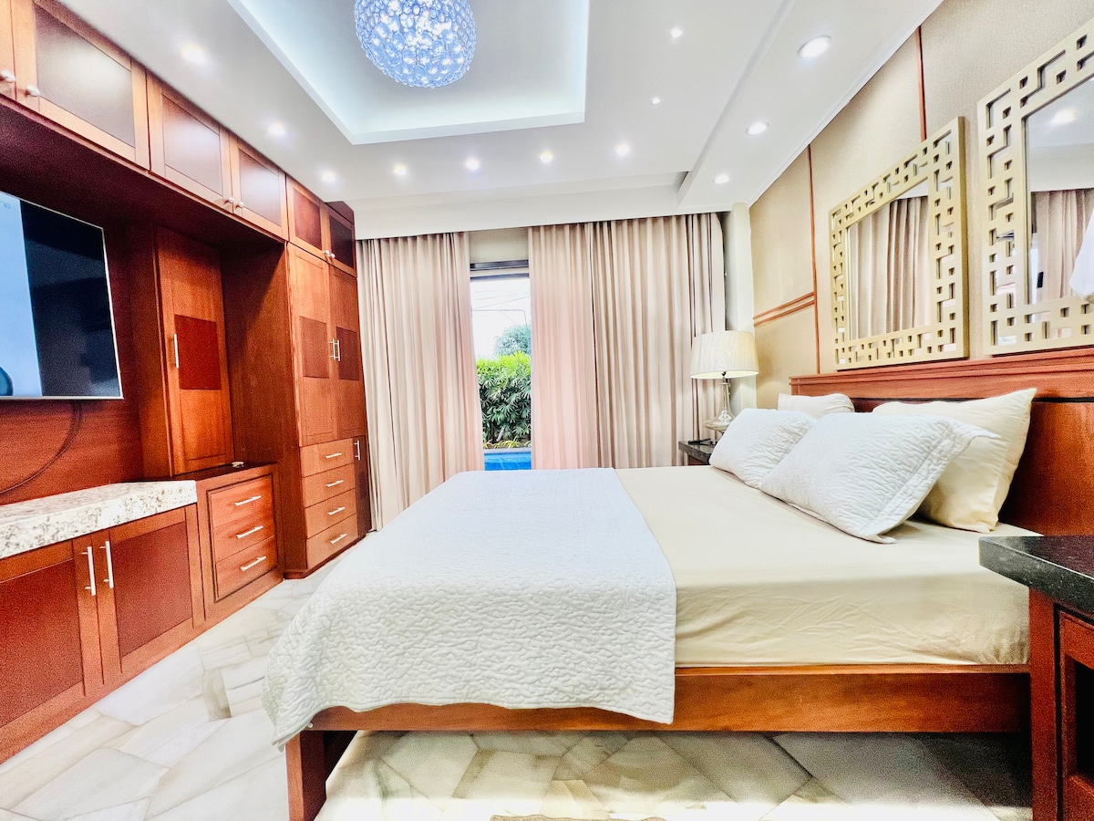 A spacious bedroom features a large wooden bed with light bedding and two pillows. Elegant cabinetry lines one wall, with a television mounted above a marble-topped dresser. Soft drapes frame a window, allowing natural light to illuminate the room. A decorative ceiling fixture adds visual interest.