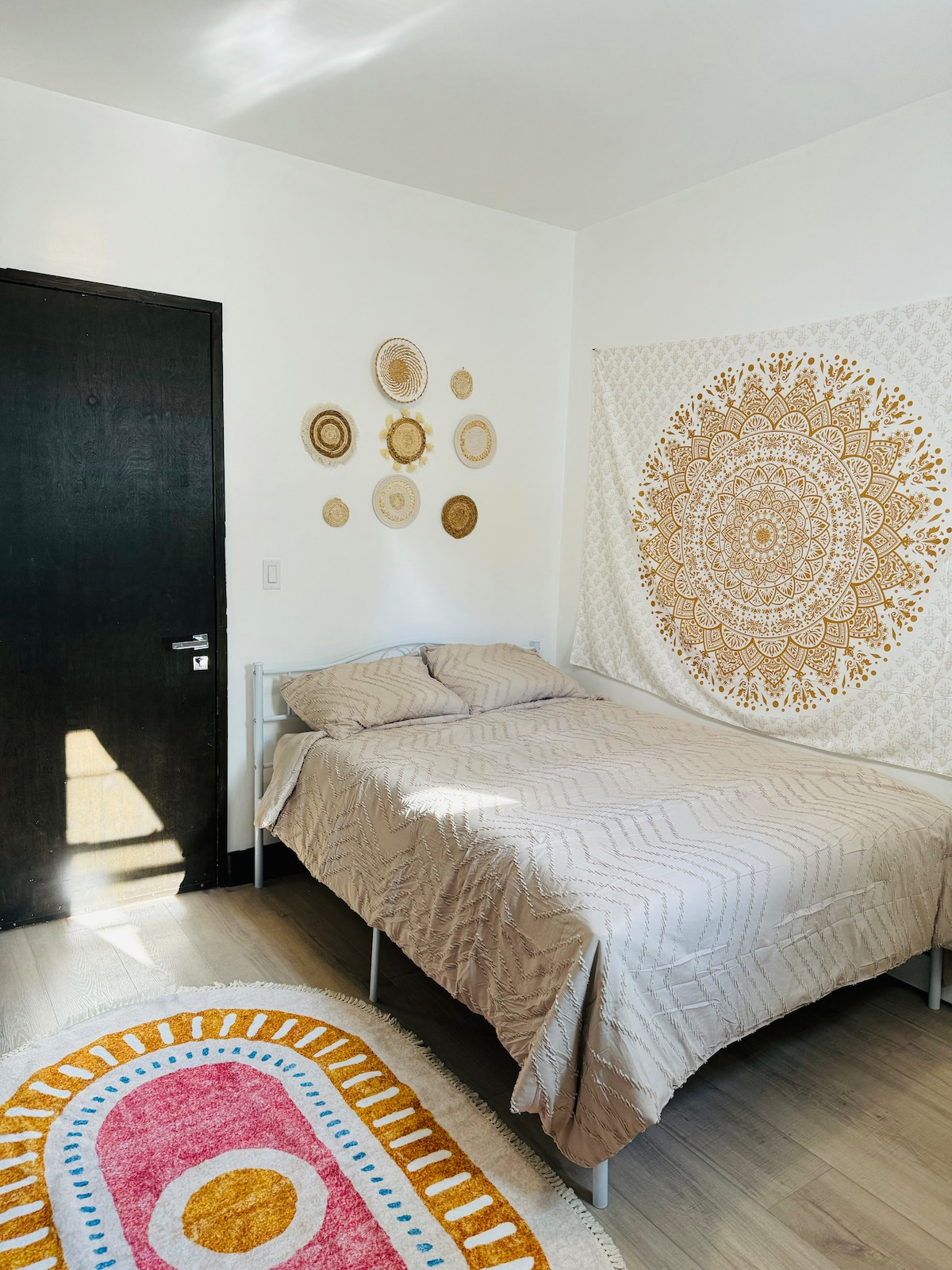 A cozy bedroom features a spacious bed dressed in neutral bedding. A decorative wall hanging in intricate patterns crowns the head of the bed. A colorful area rug with circular designs adds a touch of warmth to the room, complemented by natural light streaming through a nearby door.