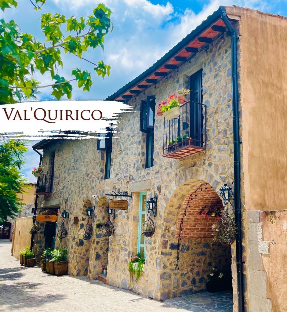 A quaint stone building features rustic architecture with arched doorways and balconies adorned with potted plants. Decorative lanterns and hanging flower baskets add charm to the inviting exterior. The surrounding pathway is lined with trees, enhancing the serene atmosphere.