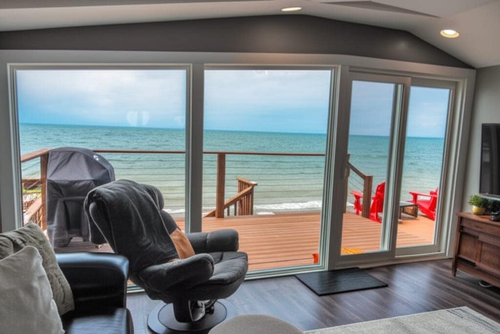 Large glass windows offer a panoramic view of Lake Erie, with the shoreline and sandy beach visible. A wooden deck features bright red outdoor chairs and a barbecue grill, enhancing the connection to the outdoor space. A comfortable black recliner is positioned nearby.