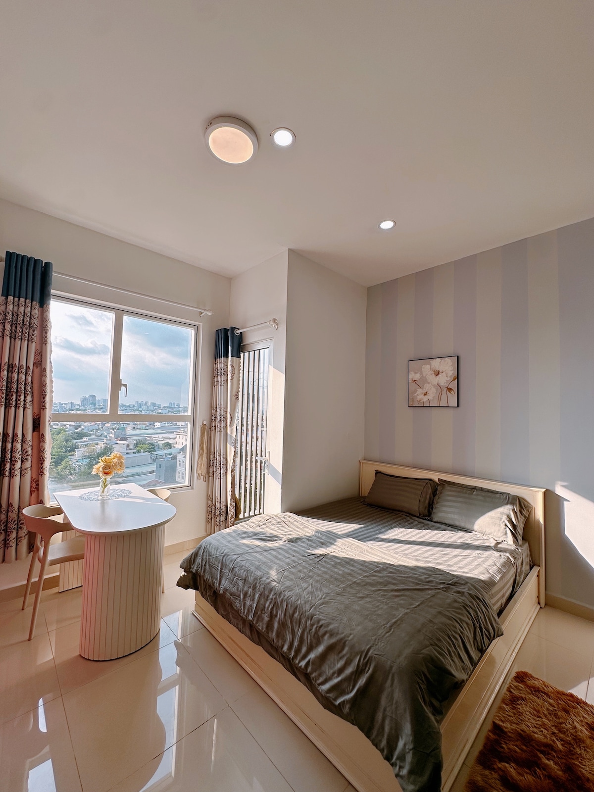 A well-lit bedroom features a spacious bed with neutral bedding and a comfortable seating area. A small round table with a minimalist design is placed beside a window, allowing natural light to fill the room. Light-colored walls contribute to a serene atmosphere.