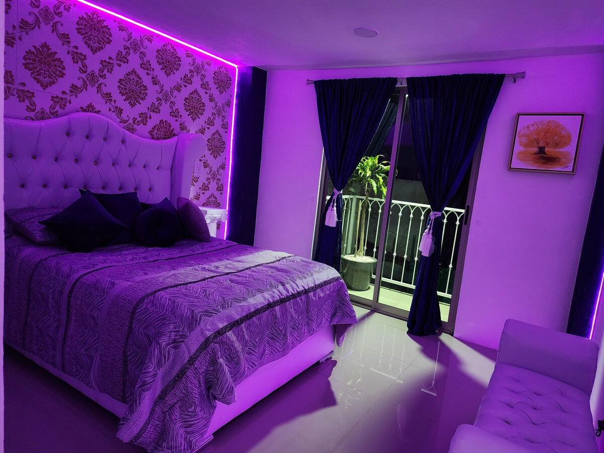 The room showcases a spacious bed with a decorative headboard, complemented by purple LED lighting along the walls. Dark curtains frame the floor-to-ceiling balcony door, leading to a private outdoor space. A classic white sofa is positioned nearby, enhancing the room's elegance.