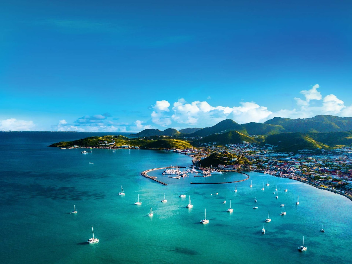 An aerial view captures the coastline dotted with sailboats, reflecting sunlight on calm waters. Lush green hills rise in the background, while the town hugs the shore, showcasing colorful buildings and a serene harbor. Soft clouds float in a clear blue sky above.
