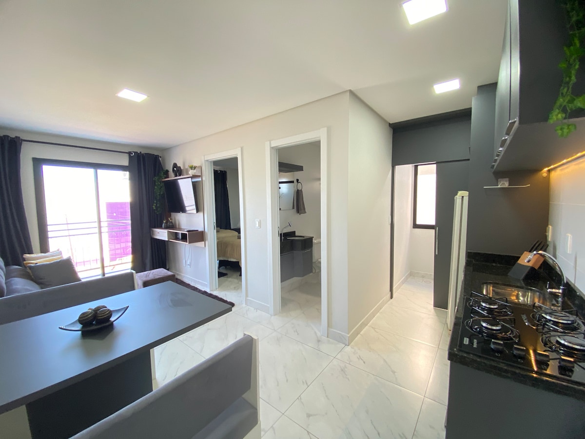 The open layout features a dining area with a table and chairs adjacent to a modern kitchen equipped with a cooktop. Natural light floods through large windows, illuminating the living space that includes a comfortable sofa and access to a private balcony.