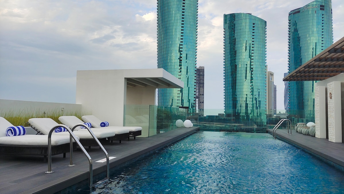 A rooftop pool area features a glistening blue pool surrounded by sleek lounge chairs adorned with blue and white towels. Towering glass skyscrapers provide a modern backdrop against the cloudy sky.