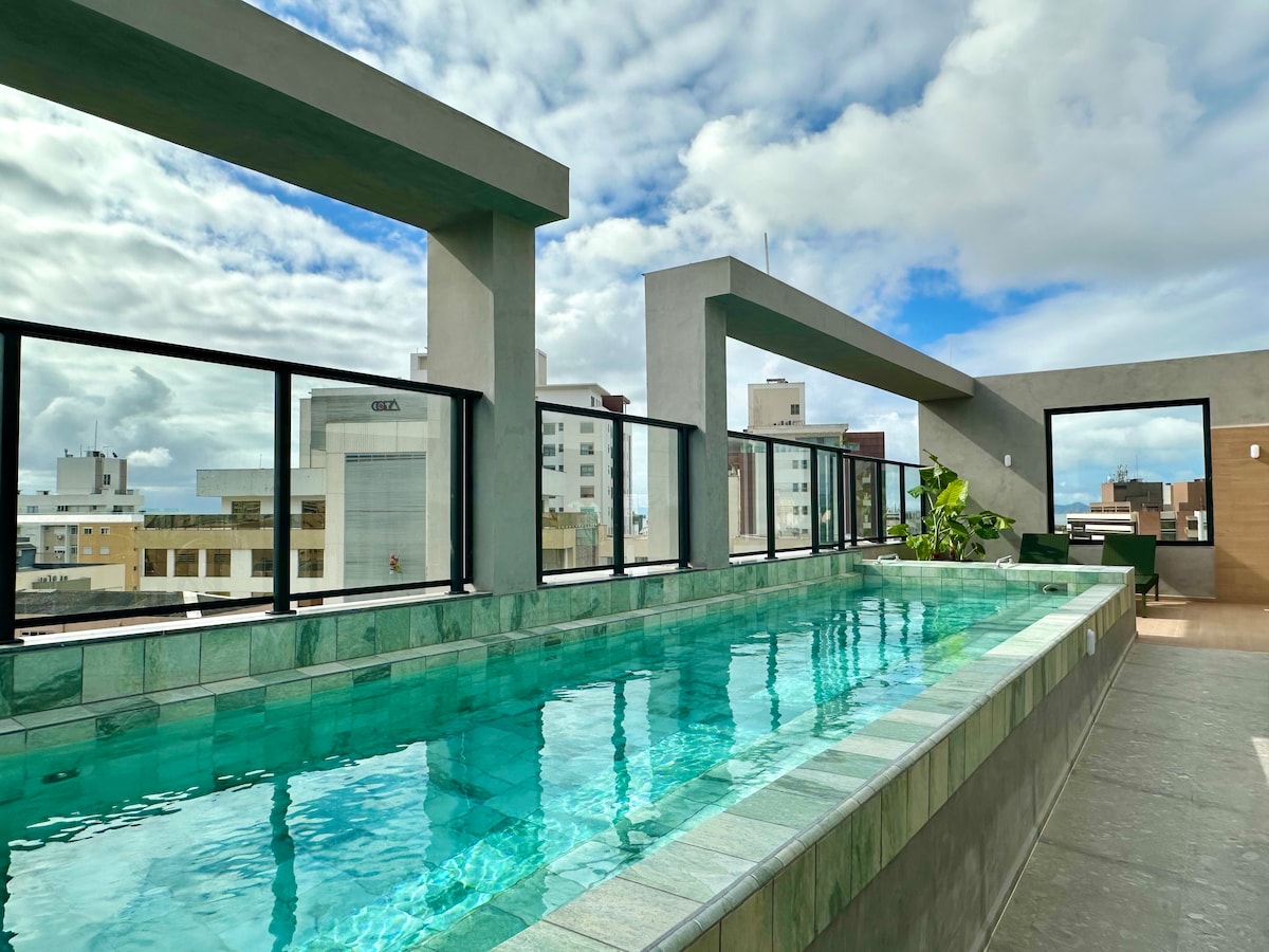 A rooftop pool is surrounded by glass railings, providing a panoramic view of the city. The water appears clear and inviting, reflecting the sky above. Planters with greenery are positioned along the edges, contributing to a serene environment.