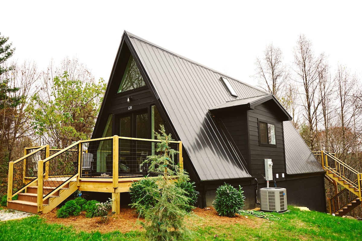 The A-frame structure features a striking black exterior and a distinctive triangular roof. A wooden deck extends from the entrance, flanked by greenery and small shrubs, creating a welcoming entrance. Large windows allow natural light to illuminate the interior.