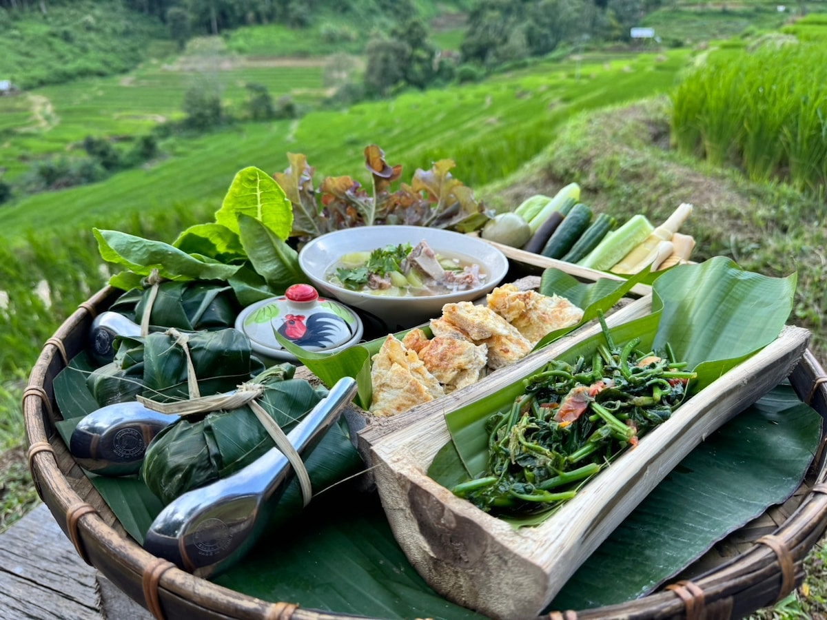 A bamboo tray filled with an assortment of local dishes is presented on a banana leaf. Fresh vegetables, a bowl of soup, and various wrapped foods create a colorful arrangement against the backdrop of green terraced fields.