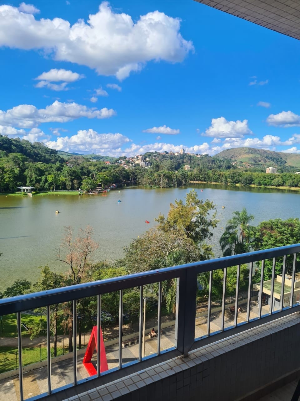 A scenic view of a shimmering lake surrounded by lush greenery is showcased. In the background, hills rise gently under a vibrant blue sky, dotted with fluffy white clouds. The image captures a peaceful atmosphere, inviting connection with nature.