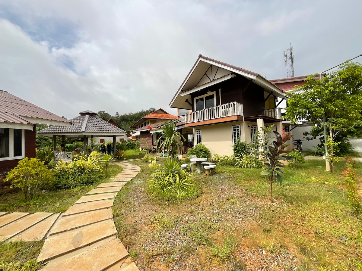 A serene outdoor area is framed by various greenery, featuring a winding stone pathway that leads to several buildings. A gazebo is positioned to one side, while comfortable seating arrangements are visible on the lawn, inviting relaxation in a tranquil setting.
