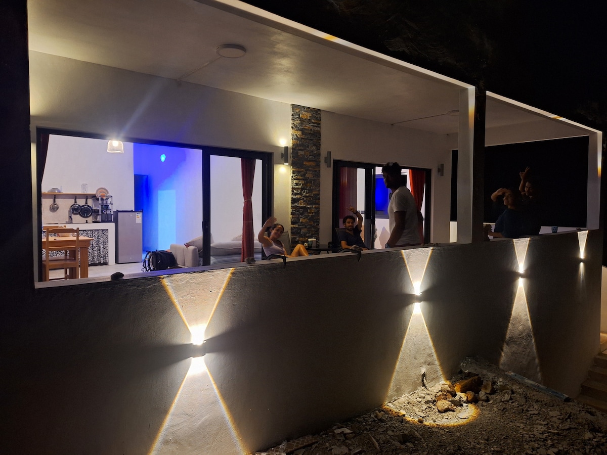 The exterior of a modern house features soft, ambient lighting illuminating the walls. Large windows offer a glimpse into the indoor space, showcasing a well-lit living area and dining area with a wooden table. A relaxed social atmosphere is suggested by the presence of several individuals.