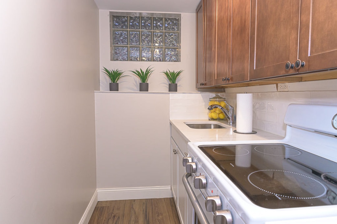 A compact kitchen features dark wood cabinetry and a modern stove. A white countertop provides workspace, while a sink is conveniently placed. Above, small potted plants are positioned against a glass block window, allowing natural light to enter the space.