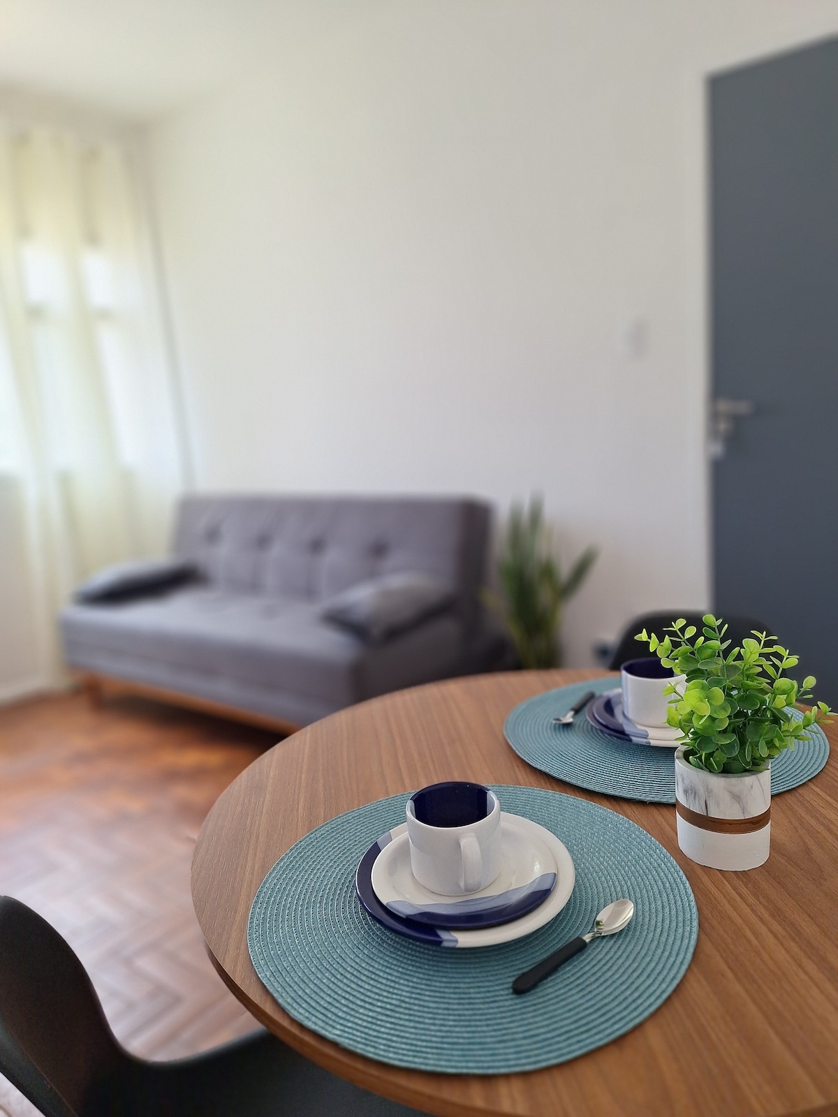 A dining table is set with plate settings and a small plant in a pot. A plush gray sofa is positioned in the background, adding comfort to the living area. Natural light enters through sheer curtains, illuminating the space.