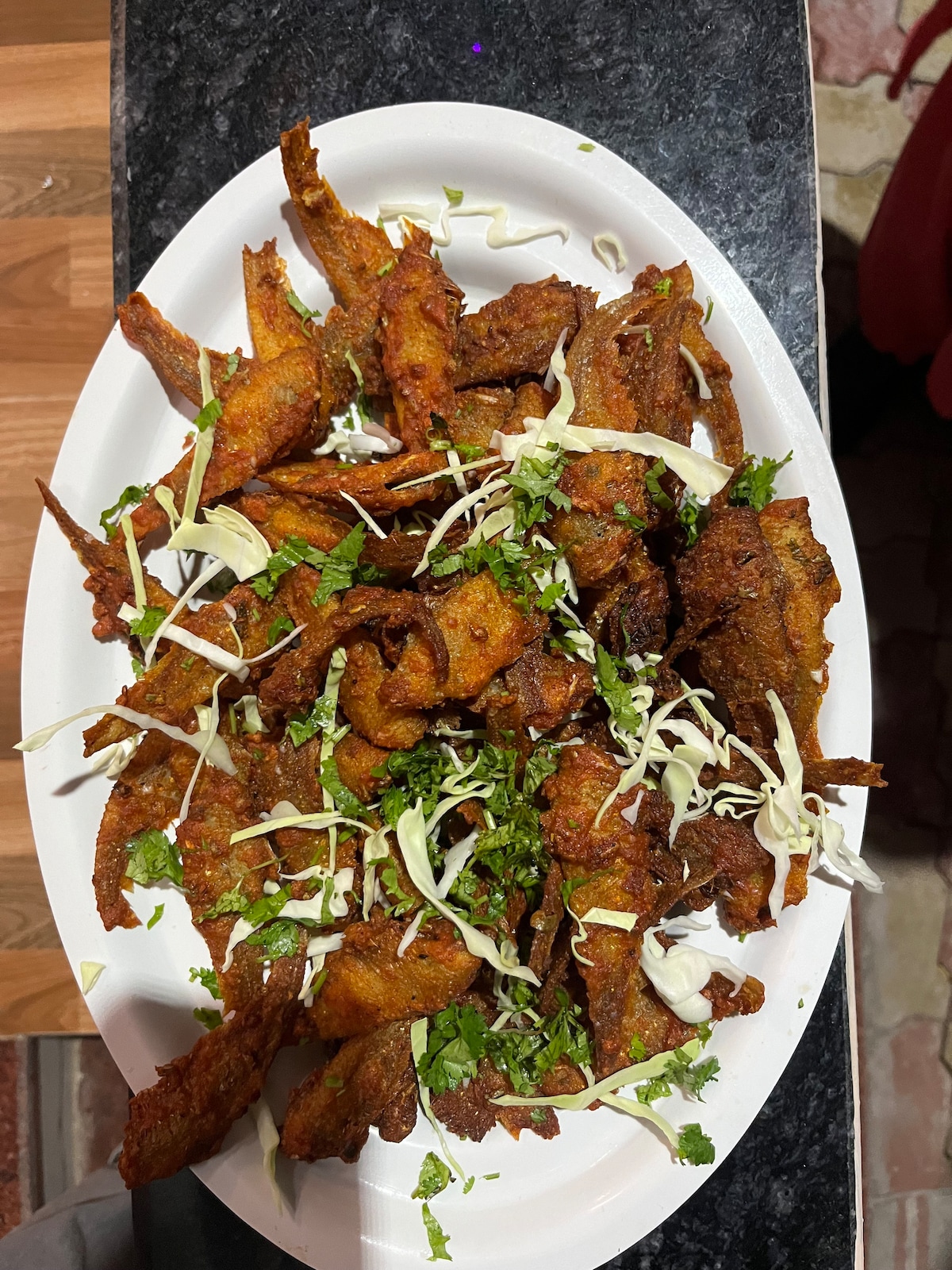 A large white platter holds an assortment of fried pieces of fish, garnished with fresh cilantro and finely shredded cabbage. The dish is presented on a dark countertop, emphasizing the golden-brown color of the fish.