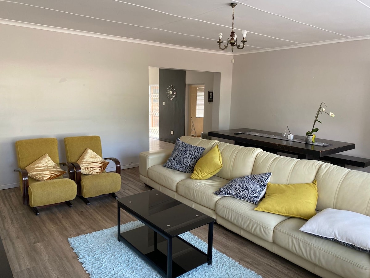 A spacious living area features a light-colored leather sofa adorned with yellow and patterned cushions. Two retro armchairs are positioned nearby. A glass coffee table is centered on a soft area rug, while a dining area is visible in the background.