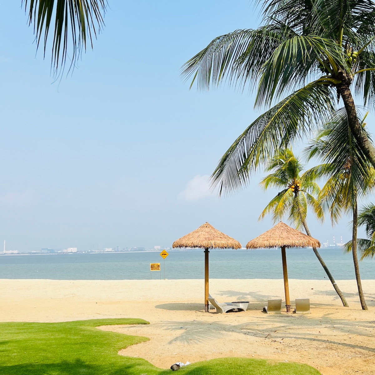 Two thatched-roof gazebos are positioned on a sandy beach, surrounded by lush greenery. Palm trees gently sway in the breeze, while the calm water extends to the horizon under a clear blue sky. A warning sign is visible near the shoreline.