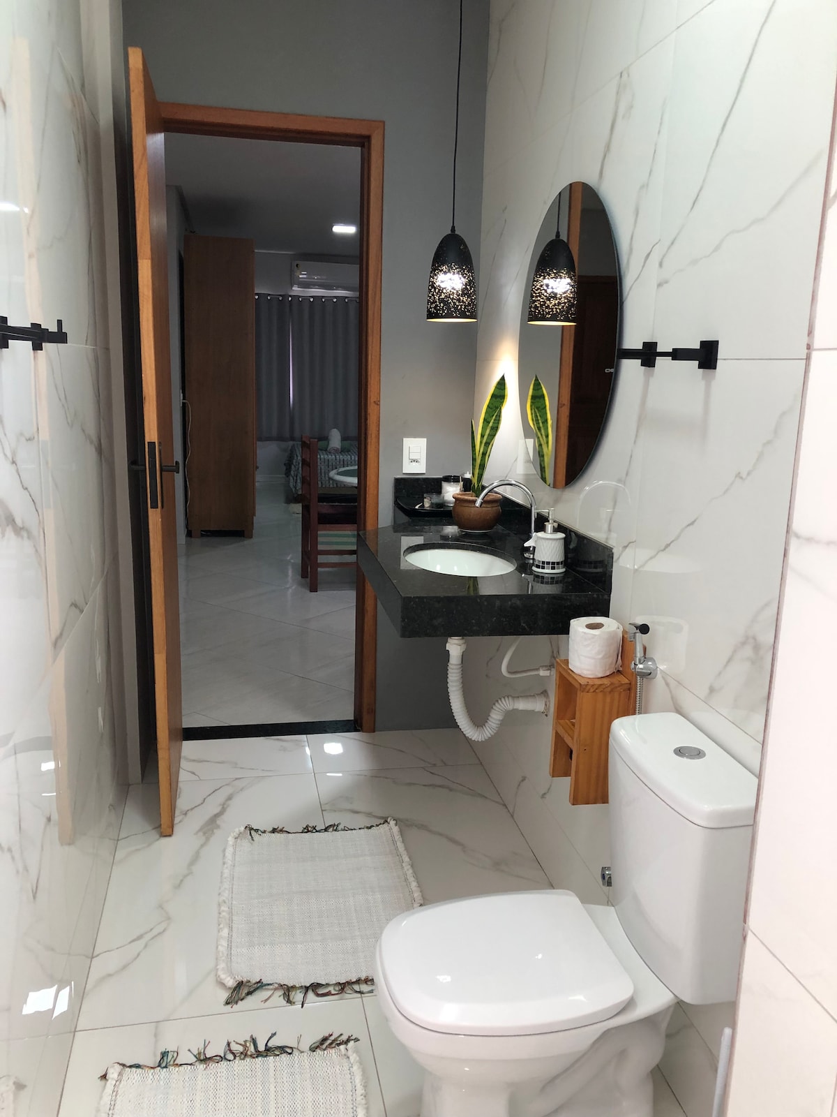 The bathroom features sleek marble walls and a modern vanity with a circular mirror. A small wooden shelf holds toiletries, while a potted plant adds a touch of greenery. Soft, neutral rugs are laid on the floor, enhancing the overall warmth of the space.