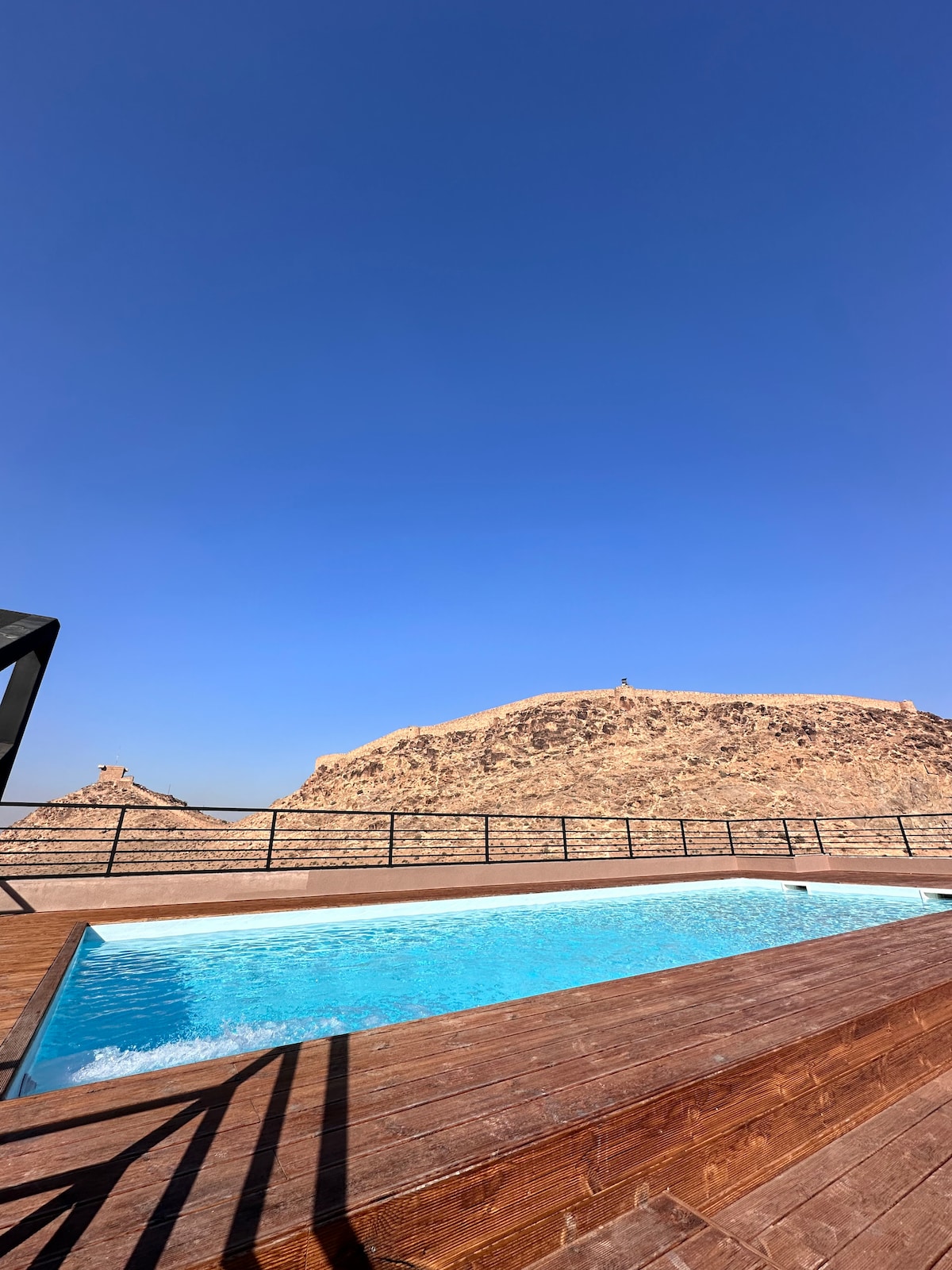 A rooftop pool is framed by a wooden deck, reflecting clear blue waters under a bright sky. A distant rocky mountain is visible in the background, providing a natural contrast to the pool's inviting design.