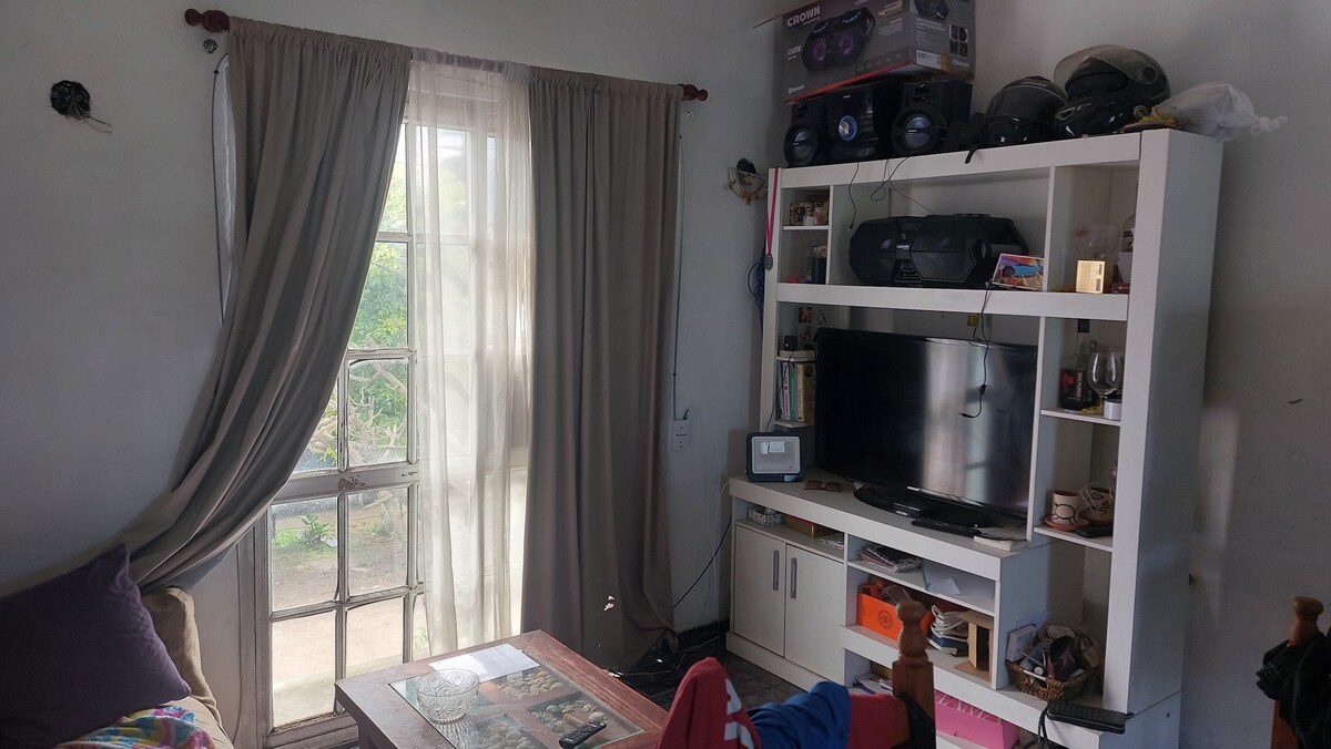 A living area is depicted featuring a white entertainment unit with a television. Various decorative items are arranged on the shelves. Light filters through a large window, with light curtains partially drawn. A cozy sofa with colorful pillows adds comfort to the space.