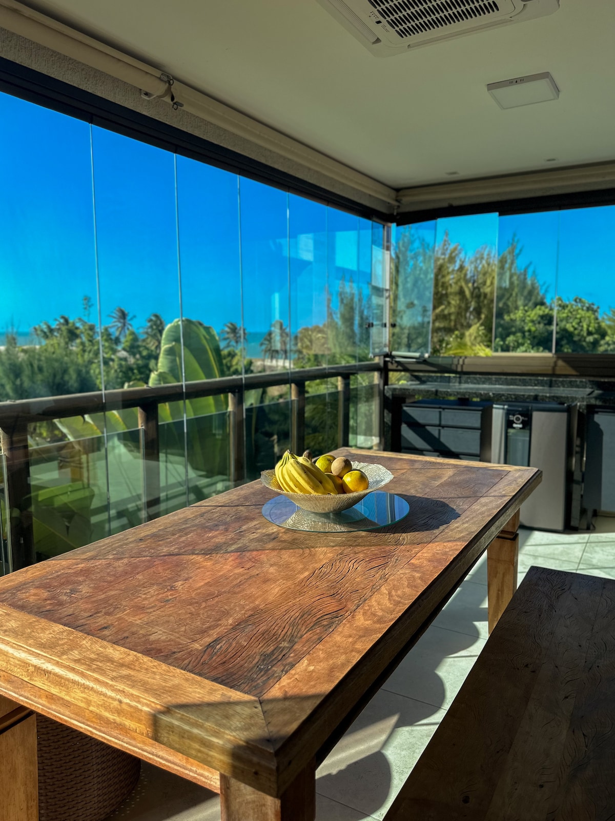 A wooden dining table is positioned in a sunlit area, showcasing a bowl of bananas and lemons. Glass panels provide an unobstructed view of the greenery outside, enhancing the connection with nature. The space is complemented by a nearby kitchenette.