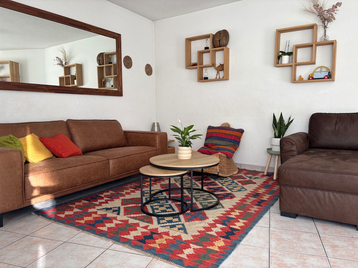 The living area features a brown couch and a matching armchair, arranged around a circular coffee table. A patterned rug covers the tiled floor, while decorative shelves display small plants and items. A large mirror reflects the room's warmth and light.