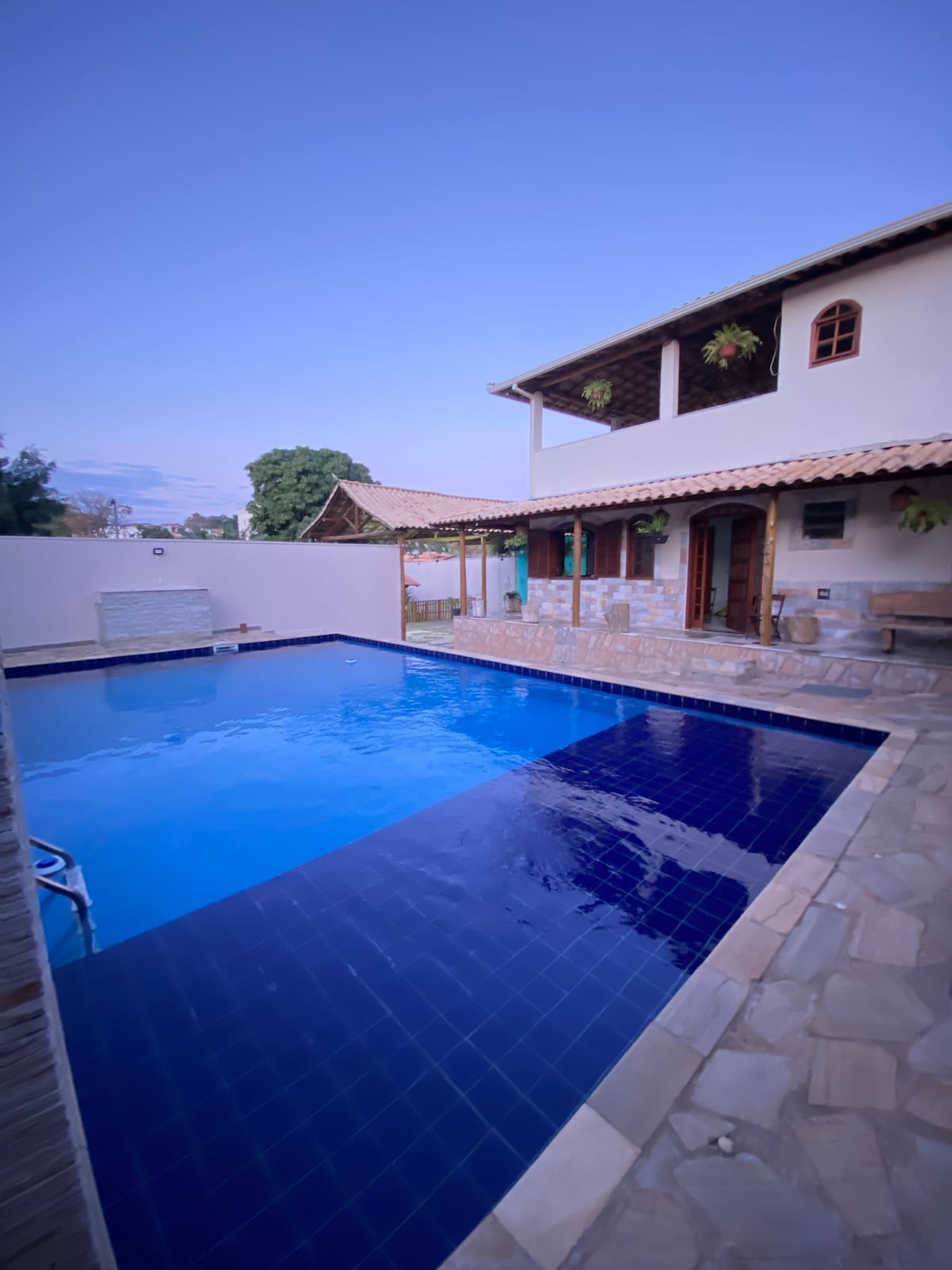 A spacious outdoor area features a large swimming pool with a deep blue surface, surrounded by stone paving. The two-story house displays a traditional design with wooden accents and large windows, offering an inviting space for relaxation and gathering.