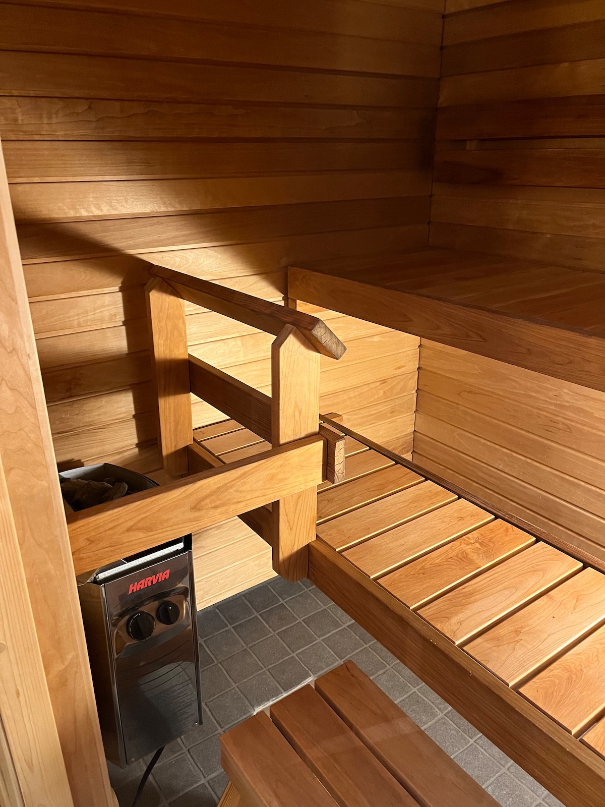 A wooden sauna interior features benches made of slatted wood, providing ample space for relaxation. The dim lighting offers a warm ambiance, while a compact sauna heater is positioned against one wall. The floor is tiled, adding to the sauna's functional design.