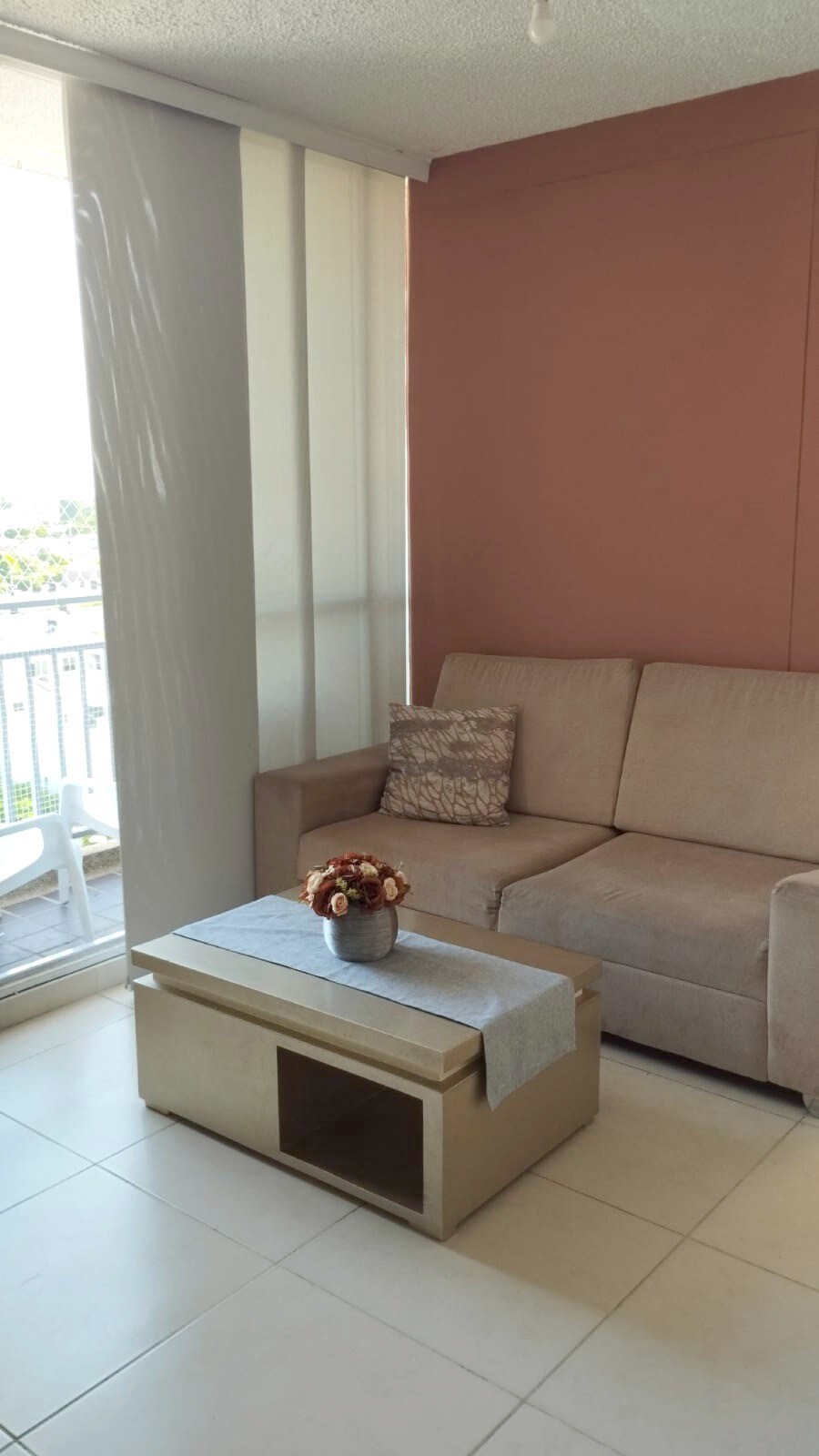 A light-filled seating area features a beige sofa adorned with a decorative pillow. A small coffee table holds a vase with dried flowers. Sheer curtains gently filter natural light, while a sliding glass door provides access to a balcony.