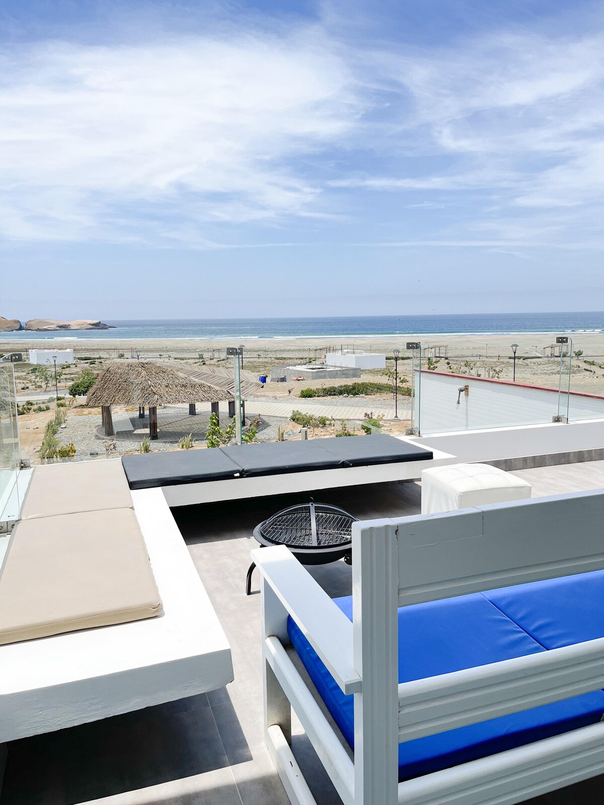 An inviting terrace features a spacious seating area with white and blue lounge furniture. A small grilling station is visible alongside a sunbed. The panoramic view of the natural beach and ocean stretches into the horizon, with shaded palapas in the background.
