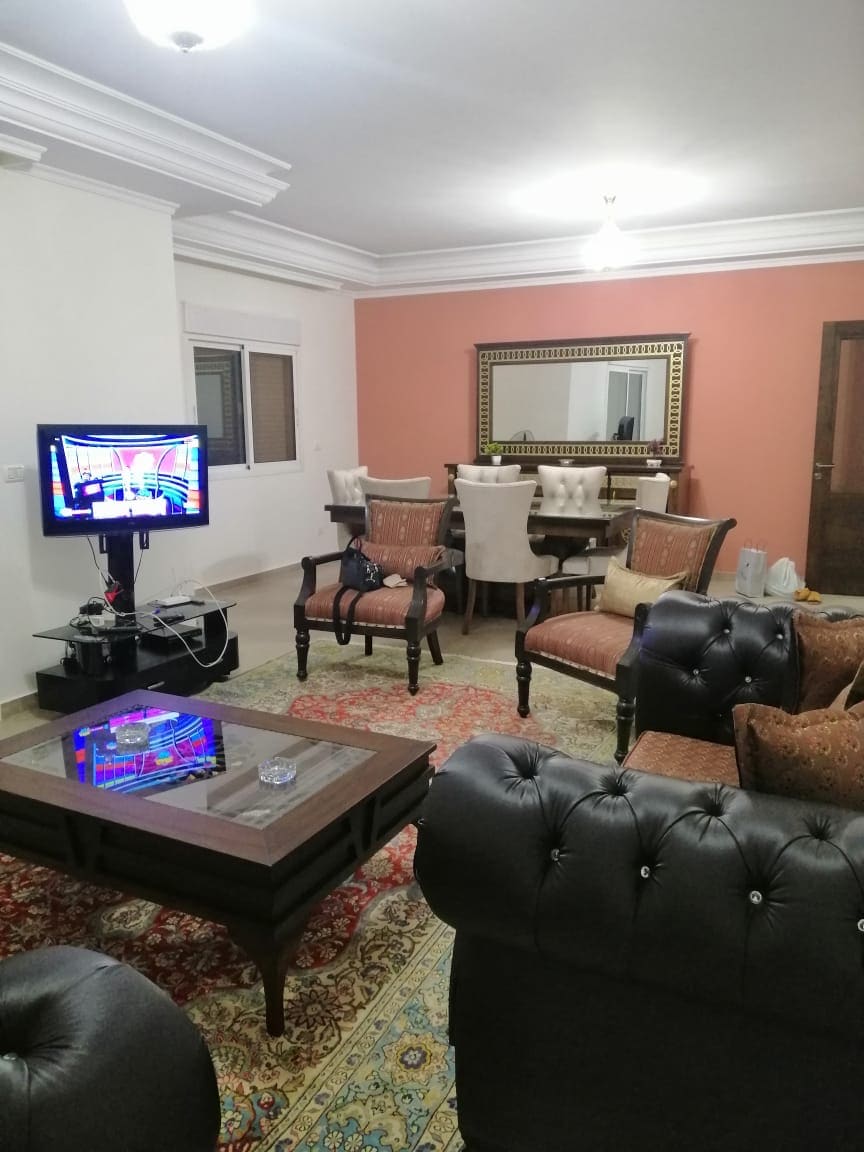 The living area features a mix of comfortable seating, including two dark leather armchairs and a plush sofa. A coffee table sits in front of the seating arrangement, while a dining table with six chairs is visible in the background. A large mirror enhances the room's depth.