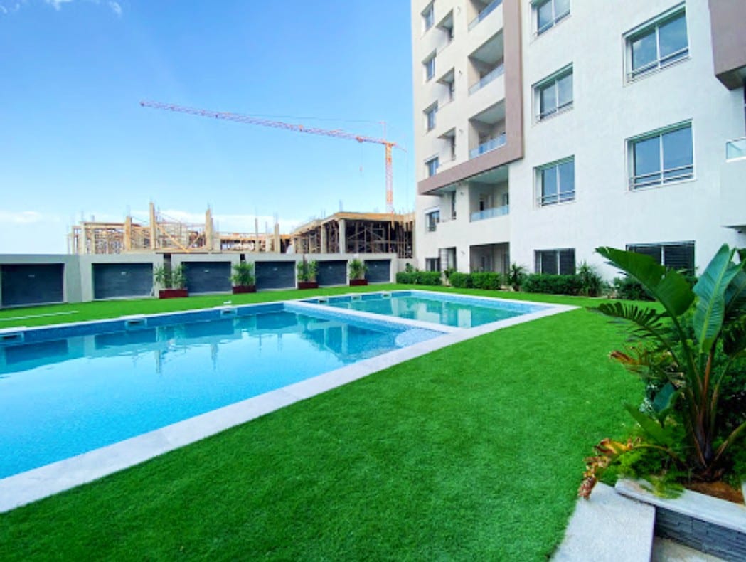 Two private swimming pools are surrounded by well-maintained green grass. The pools feature clear blue water, contrasting with the surrounding modern building. Tropical plants enhance the area, and a construction site is visible in the background, indicating ongoing development.