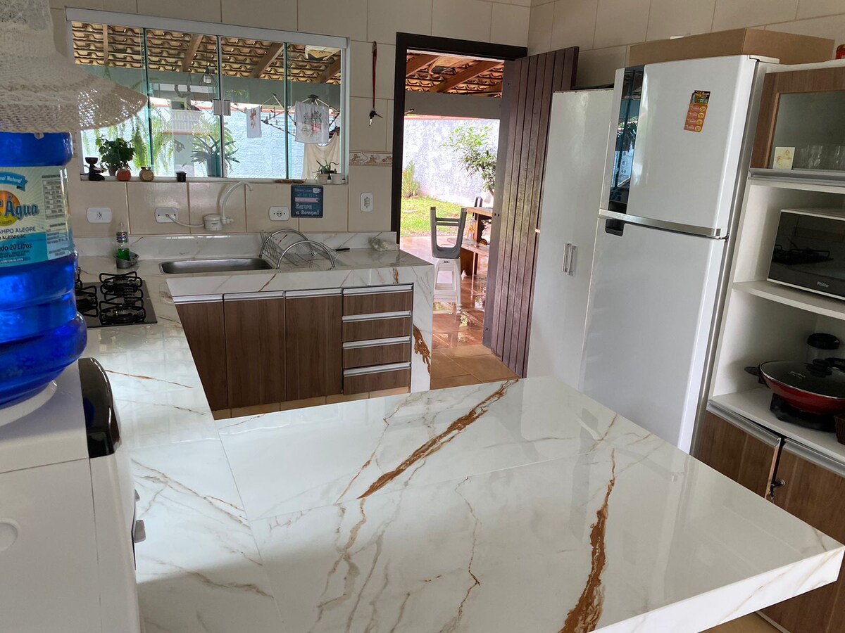 The kitchen features a spacious countertop with a marble finish and modern cabinetry. A large refrigerator is positioned adjacent to the sink area. Natural light enters through windows, illuminating the open layout that connects to an outdoor space.