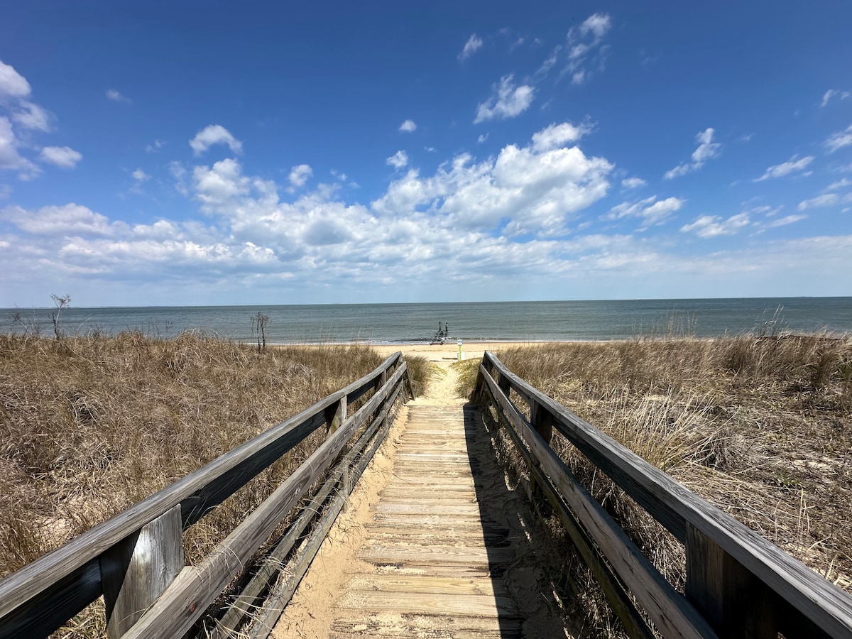 A wooden boardwalk leads directly to the beach, bordered by natural grasses on either side. The calm sea stretches under a bright blue sky with scattered clouds, creating a peaceful coastal scene. The distant shoreline is visible, inviting a sense of tranquility.