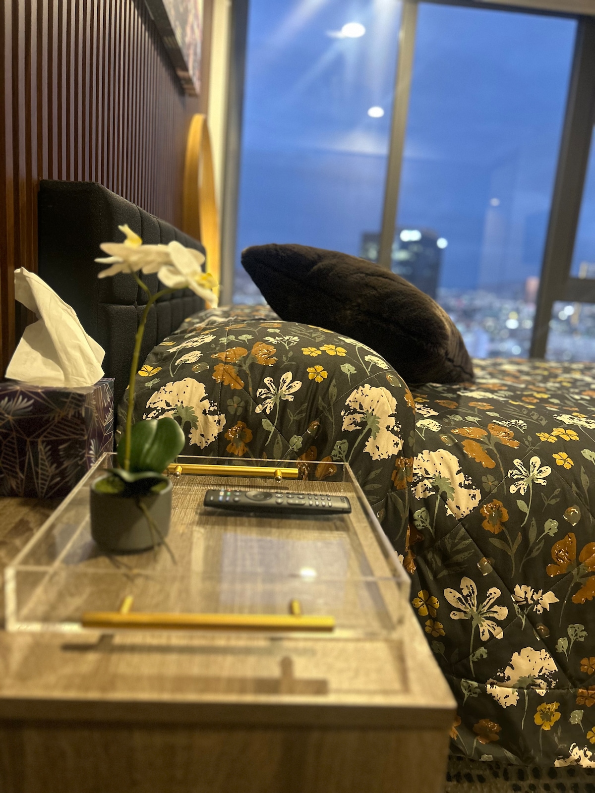 A bedside view captures a cozy sleeping area featuring a floral-patterned comforter. A small potted orchid and a tissue box adorn the nightstand, while a remote control and two yellow pencils rest on the clear surface. City lights are visible through the large window.